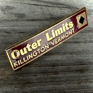May include: A rectangular metal pin with a maroon enamel background and gold-colored lettering that reads Outer Limits KILLINGTON VERMONT. The pin features a gold-bordered black diamond symbol on the right side and a gold-colored metal edge.