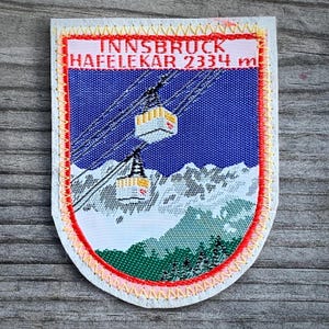 May include: A white, oval-shaped patch with red and yellow stitching around the edges. The patch features a blue background with a white mountain scene and two yellow cable cars. The text "INNSBRUCK HAFELEKAR 2334 m" is printed in black at the top of the patch.