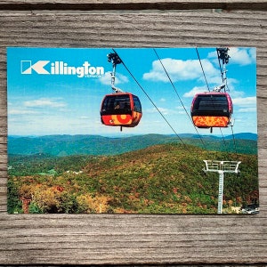 May include: A postcard featuring a view of Killington, Vermont, with two red and orange gondola cars in the sky. The postcard has the text "Killington, Vermont" in the top left corner.