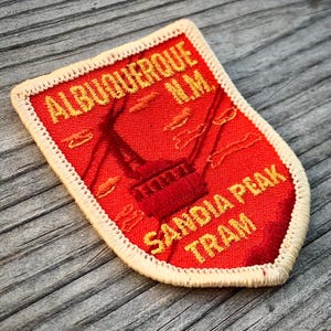 May include: Embroidered patch with the text "ALBUQUERQUE N.M. SANDIA PEAK TRAM" in yellow on a red background. The patch has a cream-colored border and features an illustration of a tramway.