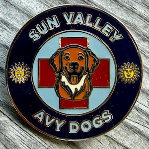 May include: A circular metal pin featuring a golden dog head centered over a red cross on a light blue background. The dark blue border displays the text SUN VALLEY at the top and AVY DOGS at the bottom, with a small yellow sun icon on each side.