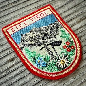 May include: Embroidered patch with a red border and the text "Zirl Tirol." The patch features a mountain scene with a blue sky, a pickaxe, and various flowers in green, red, and white. The patch is on a textured wooden surface.