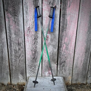 May include: A pair of green and black ski poles with blue straps and black handles. The poles are crossed and resting on a wooden surface.