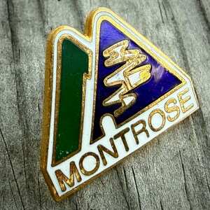May include: A vintage Montrose enamel pin with a gold border. The pin features a green and blue mountain design with the word "MONTROSE" in gold lettering. The pin is set against a weathered wood surface.