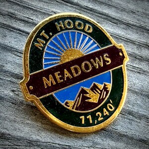 May include: A round gold-tone metal pin featuring a dark green border with the text MT. HOOD at the top and 11,240' at the bottom. The center displays a blue sky with a gold sun, a dark red horizontal banner reading MEADOWS, and mountain peaks.