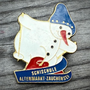 May include: A vintage enamel pin featuring a snowman with a blue hat and red scarf, skiing on blue skis. The pin has gold trim and the text "SCHISCHULE ALTENMARKT-ZAUCHENSEE" on a blue banner. The snowman has a red nose and black button eyes.