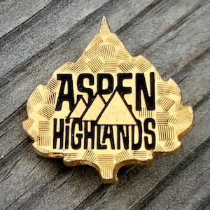 May include: A gold-colored pin in the shape of a stylized mountain range with the text "Aspen Highlands" in black lettering.