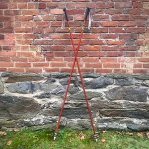 May include: A pair of vintage wooden ski poles with black tips and leather straps. The poles are crossed and leaning against a brick wall.