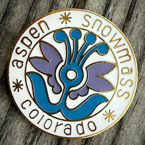 May include: A round, white enamel pin with gold trim. The pin features the words "Aspen Snowmass Colorado" in a circular pattern. The center design is a blue and purple floral motif.