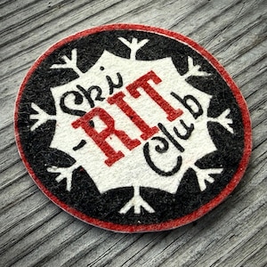 May include: Circular patch with the text "Ski RIT Club" in red and black. The patch has a white center with a snowflake design and a red border. The background is a weathered wooden surface.