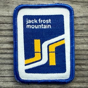 May include: A rectangular patch with a blue background and white border. The patch features the text "jack frost mountain" in white, along with yellow and white geometric shapes. The patch is likely an emblem or logo.
