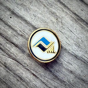 May include: A round pin with a gold border, black and white outer ring, and a white center. The center features a blue and black stylized design and the letters "ail" in gold.