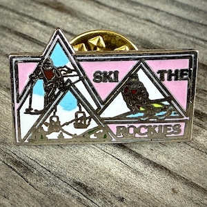 May include: A rectangular pin with a pink and white design featuring skiers and the text "SKI THE ROCKIES." The pin has a gold-colored border and a pin back. The design includes a mountain range and ski lifts.
