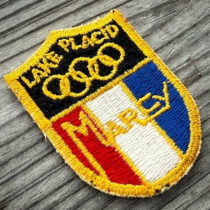 May include: Embroidered patch with a shield design. The patch features the words "Lake Placid" above the Olympic rings in gold. The shield is divided into sections of black, white, red, and blue, with the word "MARCY" in gold.