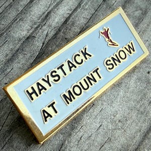 May include: A rectangular pin with a light blue background and gold trim. The pin reads "HAYSTACK AT MOUNT SNOW" in black letters. A skier graphic is in the upper right corner. The pin is likely a souvenir or collectible item.