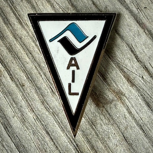 May include: A triangular pin with a black border and a white interior. The pin features a stylized design in black and teal, with the letters "A-I-L" in black. The pin is set against a weathered wooden surface.