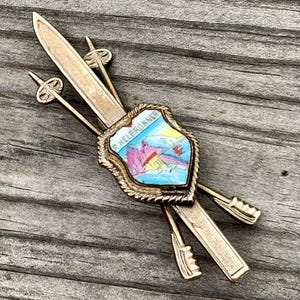 May include: A silver ski pin with a blue and white enamel shield featuring the text "P.  Felebronner" and a mountain scene. The pin is attached to two silver skis and two silver ski poles.