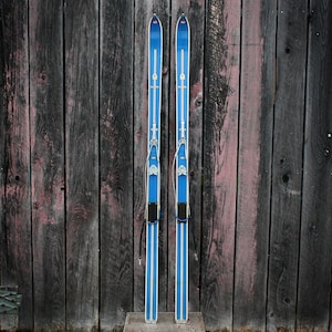 May include: A pair of vintage blue and white skis with metal bindings and a black cord attached to each ski.