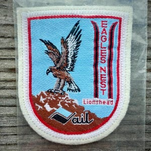 May include: Embroidered patch featuring a brown and black eagle perched on a rocky mountain. The patch has a light blue background with the words "EAGLES NEST" and "Lionshead" in red and white. The word "Vail" is also present.