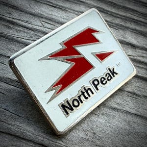 May include: A rectangular metal pin with a white enamel background featuring a stylized red lightning bolt graphic and the black text North Peak followed by a trademark symbol. The pin has a silver-toned metal border and a smooth finish.