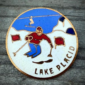 May include: A round, vintage enamel pin with a gold border. The pin features a skier in red and blue attire, skiing down a snowy slope. The words "LAKE PLACID" are written in gold lettering. A blue sky and red flags complete the design.