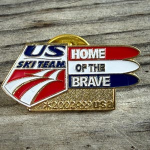 May include: A gold-toned pin featuring the text "US Ski Team" and "Home of the Brave" in red, white, and blue. The pin includes the year "2002" and the Olympic rings, with the letters "USA" at the bottom.