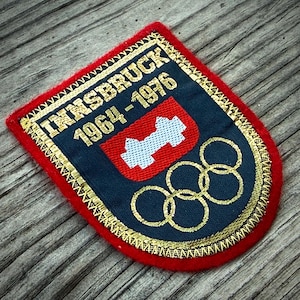 May include: Embroidered patch with the text "INNSBRUCK 1964-1976" in gold lettering. The patch features a red border, a navy blue center with a white and red emblem, and gold Olympic rings. A vintage souvenir.
