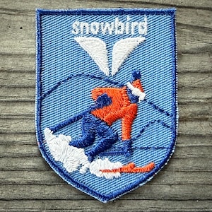 May include: Embroidered patch with a blue denim background and a shield shape. The patch features the word "snowbird" above a skier in orange and blue, with white snow and a white wing design.