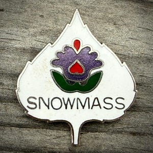 May include: A white and silver enamel pin in the shape of a leaf, featuring a purple flower with a red center and green accents. The word "SNOWMASS" is written in silver letters.
