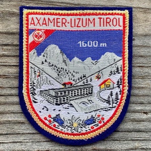 May include: Embroidered patch with the text "AXAMER-LIZUM TIROL" and "1600 m". The patch features a mountain scene with a ski lift, buildings, and trees, set against a blue sky and a red and white border.