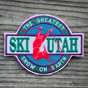 May include: Embroidered patch with the words "SKI UTAH" in green and white, with a red skier graphic. The patch is circular with a purple border and the text "THE GREATEST SNOW ON EARTH".