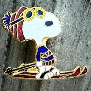 May include: A Snoopy enamel pin in a skiing pose. The pin features Snoopy wearing a blue and red ski outfit, yellow goggles, and skis. The pin has a gold-colored border and is set against a weathered wood background.