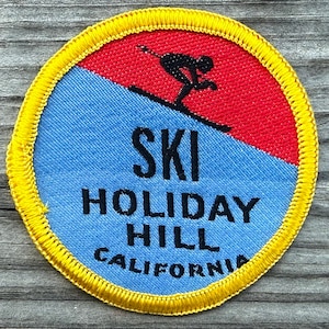May include: A circular embroidered patch with a yellow border. The top section is red with a black silhouette of a skier. The bottom section is light blue with the black text "SKI HOLIDAY HILL CALIFORNIA" printed in a bold, sans-serif font.