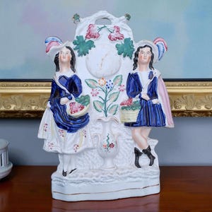 May include: A white ceramic clock with two figures in blue and white attire, holding baskets. The clock face is white with black numbers and hands. Floral and leaf accents decorate the clock's frame and base. The figures wear hats with feather accents.