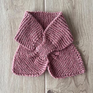 Könnte beinhalten: Ein altrosa gestrickter Schal, der auf einer hellen Holzoberfläche liegt. Der Schal ist aus dickem Garn gefertigt und weist ein deutlich sichtbares Strickmuster auf. Der Schal ist auf eine einzigartige Weise gefaltet.