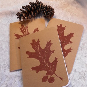 May include: Three brown paper notebooks with a red oak leaf design printed on each. The leaves are stylized and have a rustic look. The notebooks are stacked on top of each other.