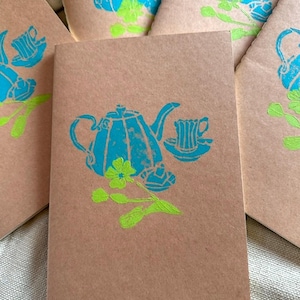 May include: A collection of rectangular notebooks with brown kraft paper covers. Each notebook features a screen-printed design in bright cyan and lime green, depicting a teapot, a teacup on a saucer, and a small floral sprig with leaves.