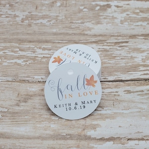 Könnte beinhalten: Ein Stapel weißer, runder Anhänger mit dem Schriftzug "fall in love" in Schreibschrift, den Namen "Keith & Mary" und dem Datum "10.6.19". Jeder Anhänger hat ein kleines Loch und eine herbstliche Blattgrafik.