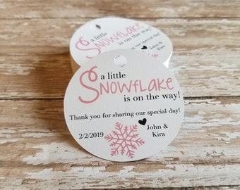 以下が含まれることがあります： ピンクの雪の結晶のデザインと「a little SNOWFLAKE is on the way!」の文字が入った白い丸いタグ。タグには日付2/2/2019とJohn & Kiraの名前も含まれています。各タグの上部には小さな穴があります。