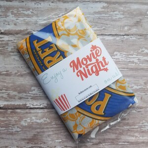 May include: A packaged bag of popcorn with a white paper band that reads "Movie Night" in red and orange. The popcorn bag has a blue and yellow design with the word "RET" visible. The band also says "Enjoy a" and "on us!"