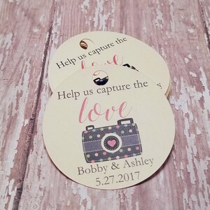 Könnte beinhalten: Runde, cremefarbene Anhänger mit dem Text "Help us capture the love" und einer Kamera-Grafik. Die Kamera ist grau mit rosa und grünen Punkten und einem rosa Herz. Die Anhänger enthalten auch die Namen "Bobby & Ashley" und das Datum "5.27.2017".