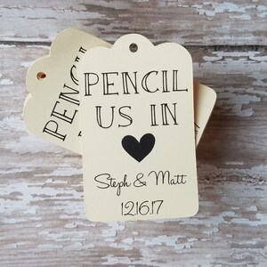 Op de afbeelding: Cr&egrave;mekleurige cadeaulabels met geschulpte randen. De labels lezen "PENCIL US IN" met een zwart hart en de namen "Steph & Matt" en de datum "12.16.17". De labels zijn gestapeld op een verweerd, lichtgekleurd houten oppervlak.
