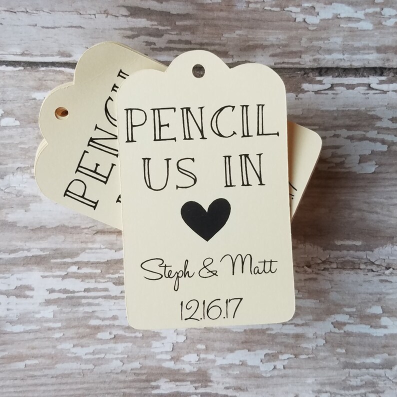 Op de afbeelding: Cr&egrave;mekleurige cadeaulabels met geschulpte randen. De labels lezen "PENCIL US IN" met een zwart hart en de namen "Steph & Matt" en de datum "12.16.17". De labels zijn gestapeld op een verweerd, lichtgekleurd houten oppervlak.
