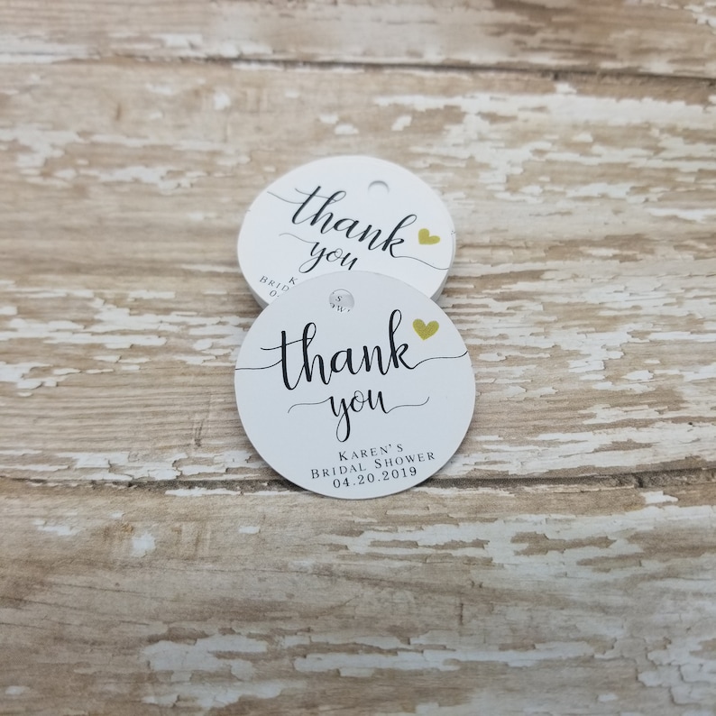 May include: Two white, round tags with the words "thank you" in elegant black script. A small gold heart is next to the word "you". The bottom of the tag reads "Karen's Bridal Shower 04.20.2019".