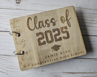 Pode incluir: Um livro de memórias de formatura em madeira com as palavras "Class of 2025" e um chapéu de formatura gravados na capa. O nome "Jamie King" e "Constantine High School" também estão gravados.