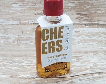 Könnte beinhalten: Kleine Flasche Zimt-Whiskey mit rotem Verschluss und weißem Anhänger. Der Anhänger trägt die Aufschrift "CHEERS to 30 years!" und den Text "Jamie's 30th Birthday July 20, 2019". Die Flasche enthält eine goldfarbene Flüssigkeit.
