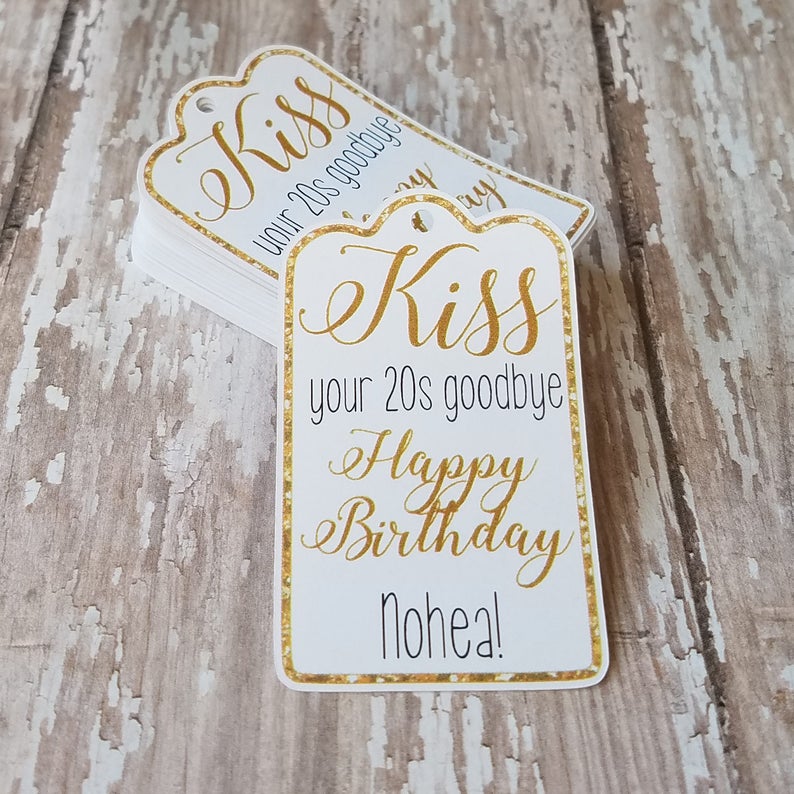 May include: White gift tags with gold text and a gold glitter border. The tags read "Kiss your 20s goodbye Happy Birthday Nohea!" in elegant script. A stack of tags is visible, suggesting a set for a birthday celebration.