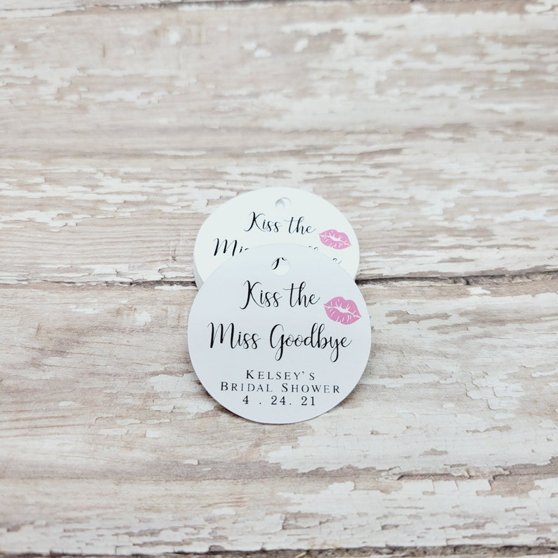 May include: Two white, round tags with the text "Kiss the Miss Goodbye" in black script, a pink lip graphic, and event details. The tags have a small hole at the top for hanging. The background is a distressed, white-painted wood surface.