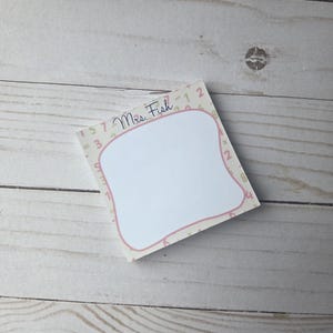 Op de afbeelding: Een vierkant notitieblok met een witte kern en een roze rand. De rand is versierd met cijfers en de naam "Mrs. Fish" in cursief. Het notitieblok ligt op een licht houten oppervlak.