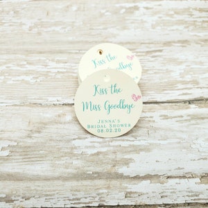 May include: A stack of round, cream-colored tags with a hole at the top. Each tag has the text "Kiss the Miss Goodbye" in teal script, with a pink glitter heart. The bottom of the tag reads "Jenna's Bridal Shower 08.02.20".